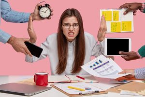 Overwhelmed office worker sitting at a desk with papers and a laptop, raising her hands in frustration while multiple hands surround her holding a clock, phone, tablet, and notepad against a pink background.